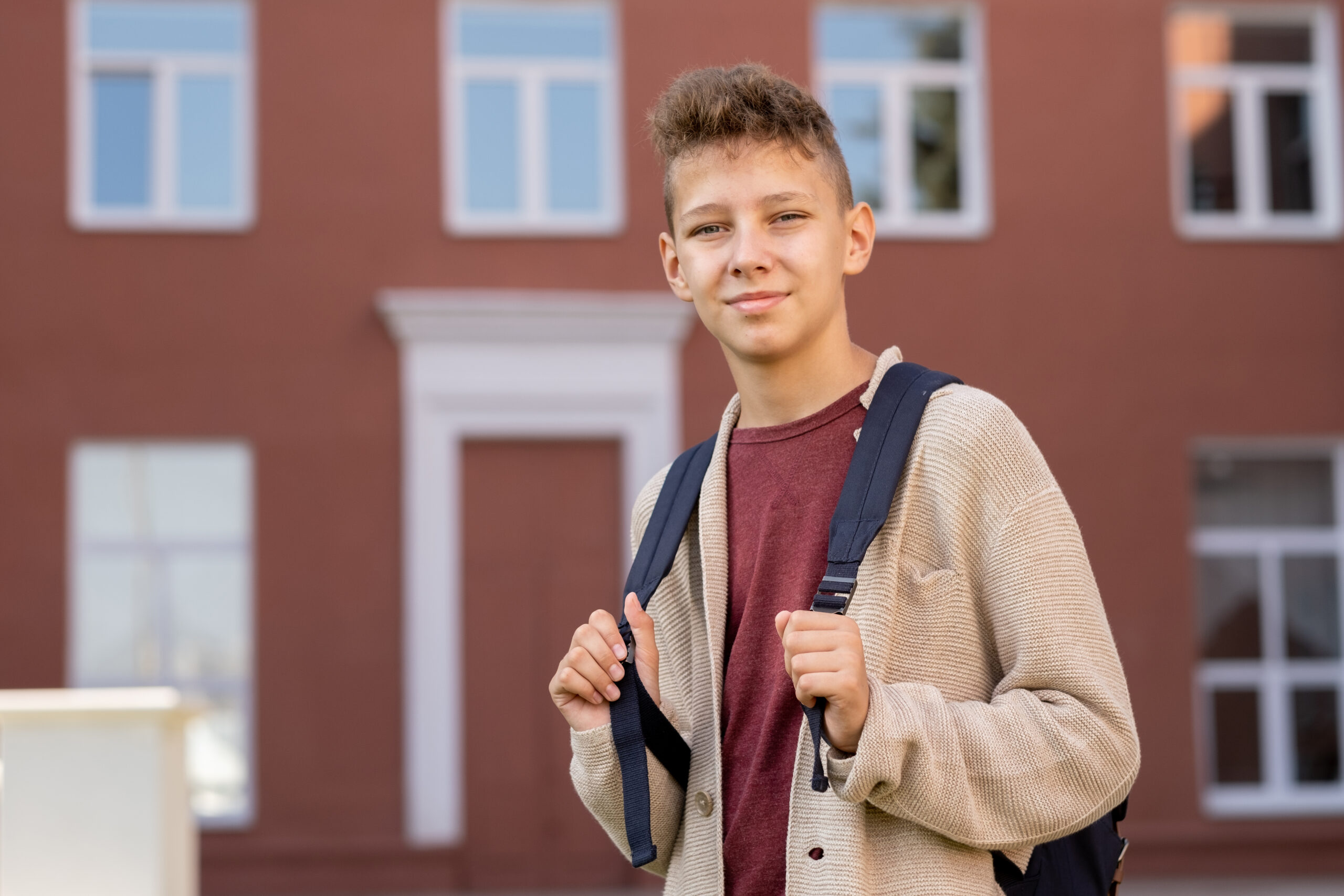 Contemporary youngster with backpack standing against building exterior and looking at you