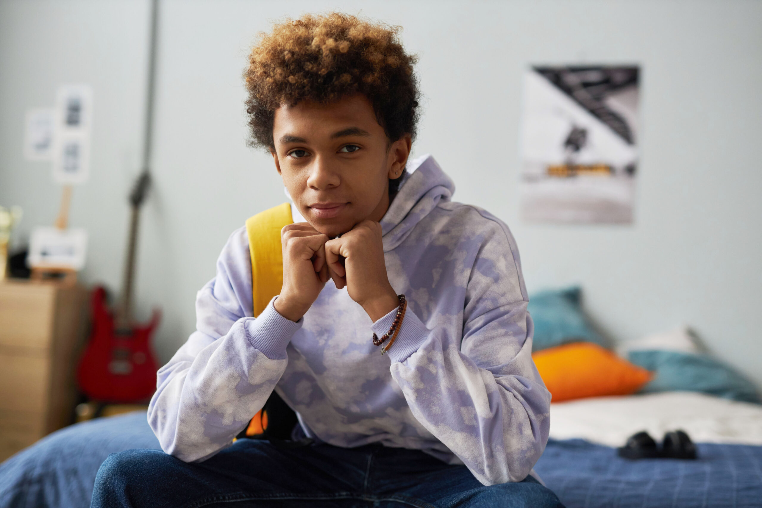 Cute serene teenage boy in casualwear keeping hands by chin while sitting on double bed in his bedroom at leisure and looking at camera